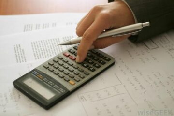 Une main tenant un stylo argenté tape sur une calculatrice. On voit aussi des papiers pleins de calculs.