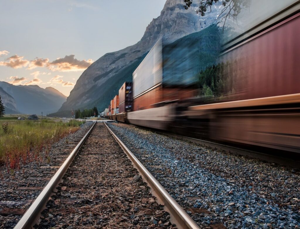 Photo d'un train de marchandises flouté, donnant une impression de vitesse. En second plan, le soleil se lève (ou se couche) derrière des montagnes.