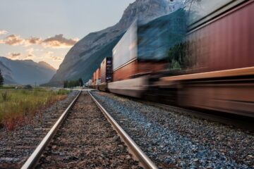 Photo d'un train de marchandises flouté, donnant une impression de vitesse. En second plan, le soleil se lève (ou se couche) derrière des montagnes.